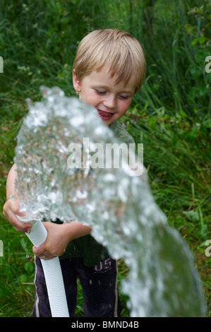 A young child boy waters garden green lawn and flowers with water from a hose pipe Stock Photo