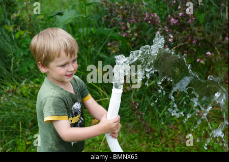 A young child boy waters garden green lawn and flowers with water from a hose pipe Stock Photo