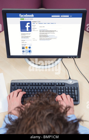 Overhead shot of a man sat at a desk working on a computer, looking at a facebook page on his screen. Stock Photo