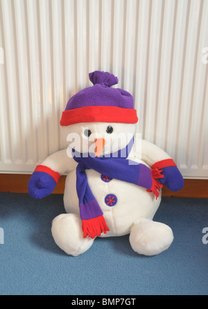 a soft cuddly toy snow man wearing a hat and scarf sits on the carpet leaning against the radiator. Stock Photo