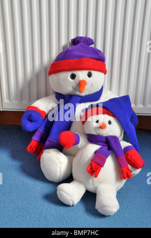 two soft toy snow men wearing hats and scarves sit on the carpet leaning against the radiator. Stock Photo
