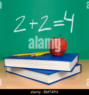 2 + 2 = 4 written on a chalkboard. Books, pencils and an apple on the foreground. Stock Photo