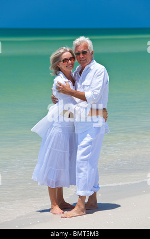 Romantic and happy senior man and woman couple dressed in white and embracing on a deserted tropical beach with blue sky Stock Photo