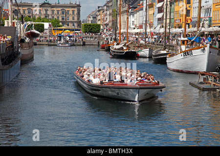 Canal tour cruise boat full of summer tourists leaving Nyhavn on a sightseeing cruise in Copenhagen passing the crowded dockside pavement restaurants. Stock Photo