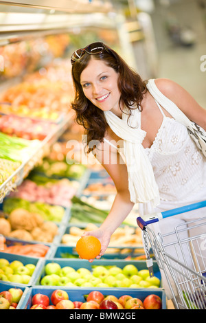 Grocery store - smiling woman shopping with trolley in supermarket, holding orange Stock Photo