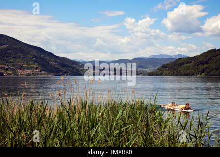 view across Lago di Caldonazzo, Trento, Italy, catttail reed and two girls paddling on surfboard in foreground Stock Photo