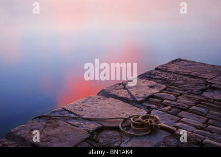 A coil of rope lying on a stone jetty next to the sea in Swanage, Dorset. The water is calm and lit by the setting sun. Stock Photo