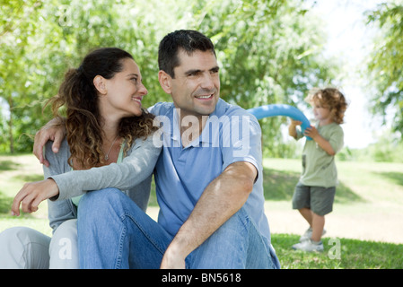 Couple relaxing together outdoors, woman looking over shoulder at son playing in background Stock Photo