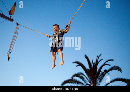 A young boy bounces high on a fairground Bungee Jump against the blue evening sky and palm trees in Alcudia, Majorca. Stock Photo