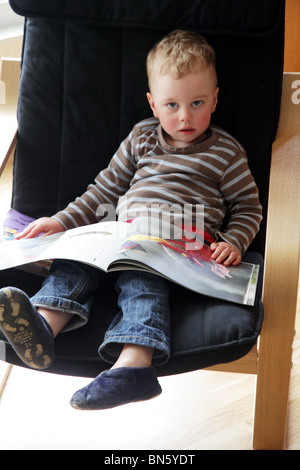 Boy toddler reading the book Room On The Broom by Julia Donaldson Axel Scheffler sat alone in a chair MODEL RELEASED Stock Photo