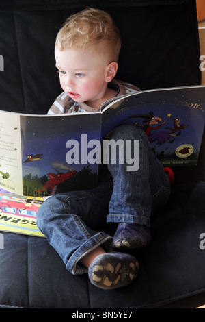 Boy toddler reading the book Room On The Broom by Julia Donaldson Axel Scheffler sat alone in a chair MODEL RELEASED Stock Photo