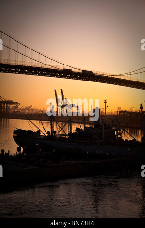 A silhouette of a ship at port, the Gerald Desmond Bridge with traffic, and industrial cranes at dawn.in the Port of Long Beach. Stock Photo