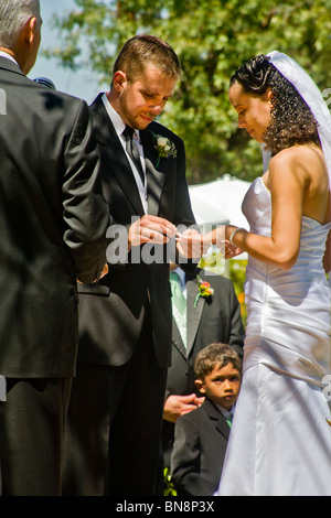 Caucasian groom puts the wedding ring on finger of his mixed-race bride at their formal outdoor wedding ceremony.  MODEL RELEASE Stock Photo