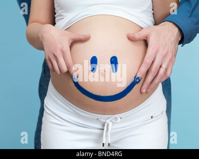 Pregnant young woman and her husband painting a happy smiley face on her belly. Stock Photo