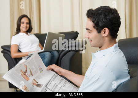 Man reading a newspaper with his wife using a laptop Stock Photo