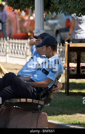 Two Turkish police (Polis) officers resting in the shade on a wooden park bench with passers by in the background in Istanbul Stock Photo