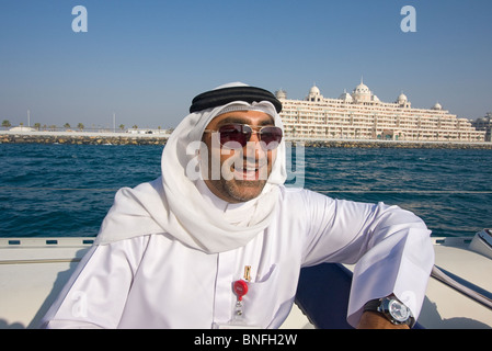 Local Arab man in traditional dress rides in tour boat past one huge hotels located on the palm shaped island complex in Dubai Stock Photo