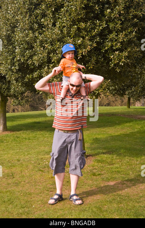 Vertical portrait of a young father carrying his young son on his shoulders in the sunshine.