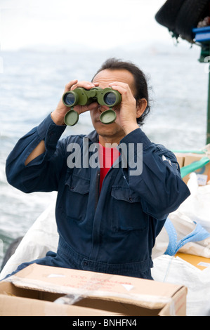 Sailor dressed in blue coverall on a small vessel looking through binoculars. Stock Photo