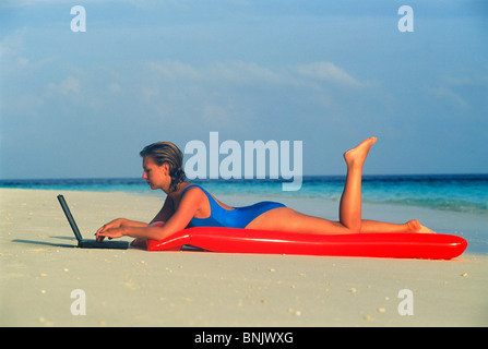 Woman laying on air mattress on sandy beach using laptop during working holiday Stock Photo