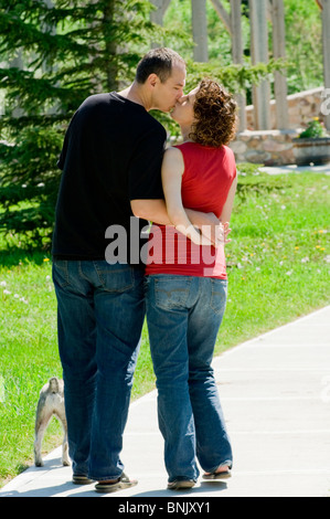 Couple kissing while walking away outside in the summer time with their dog on a walkway surrounded by lush green grass. Stock Photo