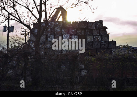 Crushed aluminum cubes are stacked high by a large yellow crane as the sun sets over a scrapyard. Stock Photo