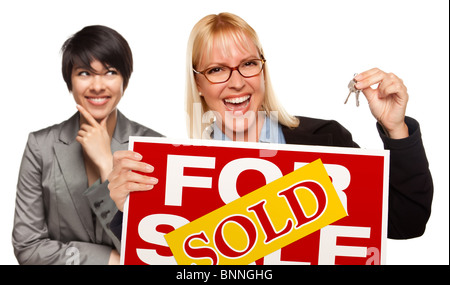 Hispanic Female Behind with Attractive Blonde in Front Holding Keys and Sold For Sale Sign Isolated on a White Background. Stock Photo