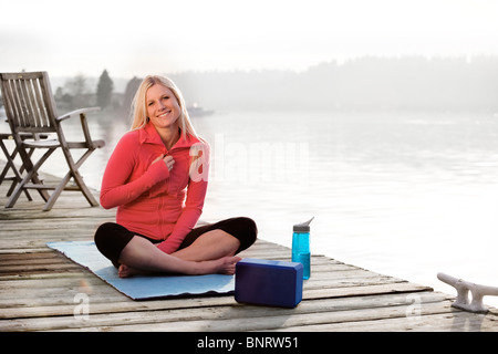 A young woman hanging out on a dock over Lake Washington and smiling at camera while sitting on a yoga mat. Stock Photo
