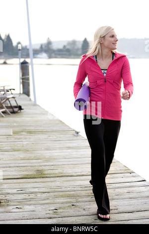 A young woman walking out on a dock over Lake Washington and carrying a rolled yoga mat. Stock Photo