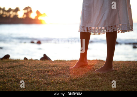 A barefoot woman in a dress walks on grass above the sea at sunset. Stock Photo