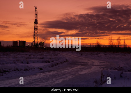 Snowy road at dusk with oil drilling rig in background Stock Photo