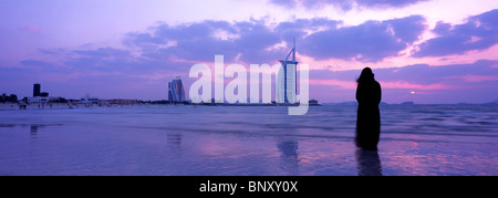 Panoramic shot of woman in traditional abaya admiring the sunset at one of Dubai's beaches with Burj Al Arab in background. Stock Photo