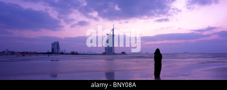 Panoramic shot of woman in traditional abaya admiring the sunset at one of Dubai's beaches with Burj Al Arab in background. Stock Photo