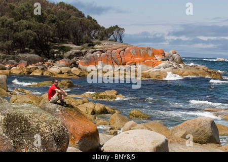Male dressed in red t-shirt and shorts sat on rock looking out to sea.  Behind can be seen rocks covered in orange lichen. Stock Photo