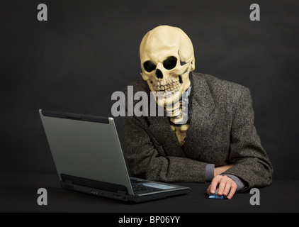 The terrible skeleton in a suit sits at black office with the laptop Stock Photo