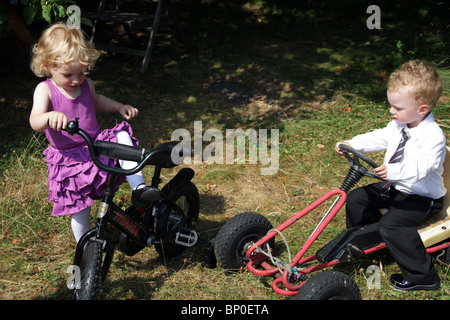 Little girl boy siblings toddlers playing together in party clothes dressed up bicycle and soapbox car kart MODEL RELEASED Stock Photo