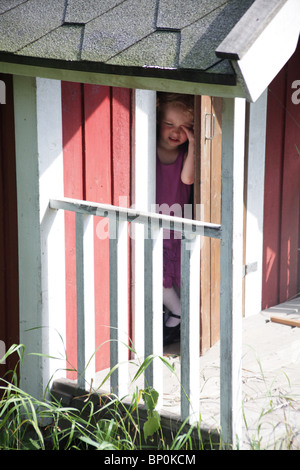 Little girl toddler in party clothes being nosy peeking out and opening an outside wooden playhouse door MODEL RELEASED Stock Photo