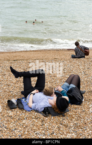 two young people relaxing on Brighton shingle beach, man playing a guitar and swimmers in the sea. Stock Photo