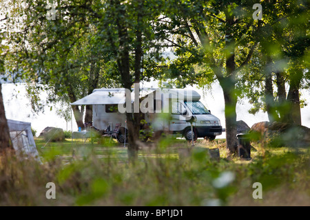 A camper parked at a lake, a man is sitting in the shadows relaxing Stock Photo