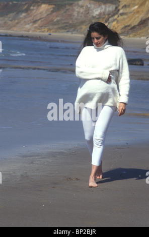 mixed race heavily pregnant woman walking on the beach warmly dressed in a white jumper Stock Photo