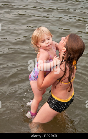 Caucasian 9 year old girl is lifting and helping a younger toddler girl in the lake as they are in bathing suits having fun.