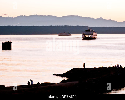 sunset watchers on rock jetty silhouetted against clear blush twilight as incoming ferry passes outgoing approaching Edmonds WA Stock Photo
