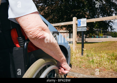 Elderly woman in a wheelchair by a car parked in blue badge disabled parking bay with sign for wheelchair users only. UK. Stock Photo