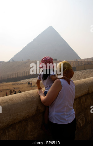 Lady tourist wearing a headscarf holds a young girl in a beanie hat against a wall looking at the pyramids of Giza Stock Photo