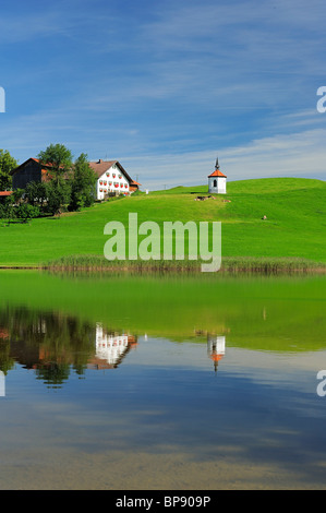 Reflection of upper Bavarian traditional farmhouse and chapel on lake, East Allgaeu, Bavaria, Germany Stock Photo