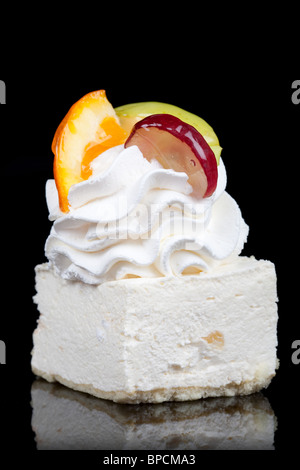 A close-up of a small, square-shaped white mousse cake topped with a generous swirl of whipped cream. The dessert is garnished with a slice of orange, a slice of green apple, and a slice of red Stock Photo