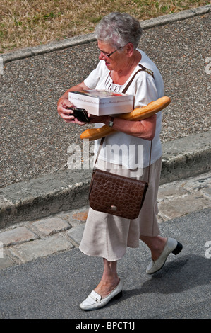 Older woman walking alongside pavement carrying crocodile hand bag, baguette and box of cakes - France. Stock Photo