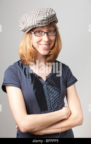 Young woman in a blue shirt and plaid flat cap, wearing glasses and smiling with arms crossed against a neutral background. Stock Photo