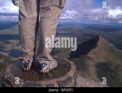 Walker standing on trig point on summit of Snowdon (Yr Wyddfa) near Llanberis Gwynedd Snowdonia National Park North Wales UK Stock Photo