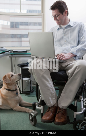 Man in a wheelchair working on a laptop with a service dog sitting near him Stock Photo
